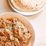 shredded chicken in a white bowl with a plate or tortillas beside the chicken