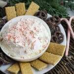 creamy white dip in a white bowl topped with crushed candy canes and surrounded by chocolate covered pretzels.