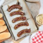 overhead photo of oven baked brats on a baking sheet with buns on the side and a bowl of sauerkraut.
