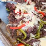 cooked sliced beer with cooked red and green peppers on a sheet pan. The sandwich filling is topped with a melted white cheese sauce. There is a spatula scooping the cheesesteak from the pan.