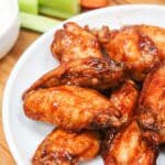 cooked wings in bbq sauce piled on a white plate. Sliced carrots and celery on the side with ranch dressing.