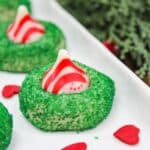 sugar cookies with small green sprinkles. A red and white kiss candy is placed in the middle. Cookies are served on a white plate.