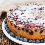 blueberry lemon cake on a white plate topped with powdered sugar and surrounded by fresh blueberries.