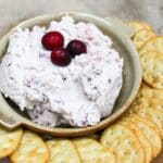 pink cranberry jalapeno dip topped with fresh cranberries on a bowl. Crackers are served on the side of the bowl.