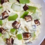 Creamy salad in a white bowl topped with chopped snickers bars and fresh green apple pieces.